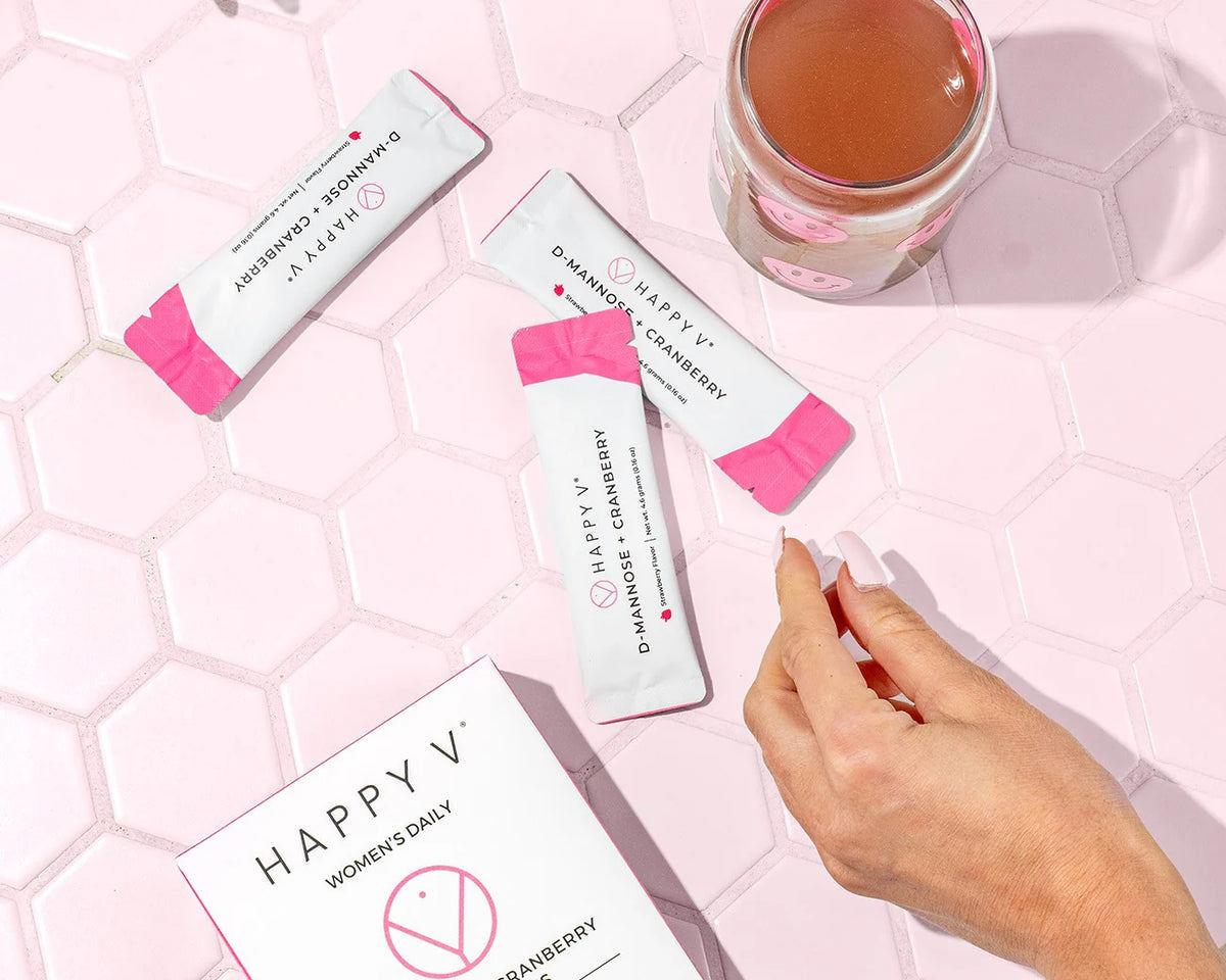 A box of Happy V's D-Mannose and Cranberry stick packs with some stick packs laying next to a glass of diluted Cranberry and D-mannose supplements. 
