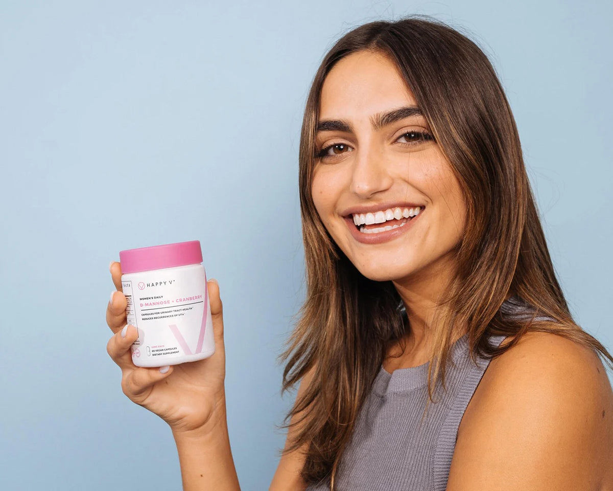 A smiling woman looking at the camera and holding a bottle of Happy V's D-Mannose and Cranberry supplements.