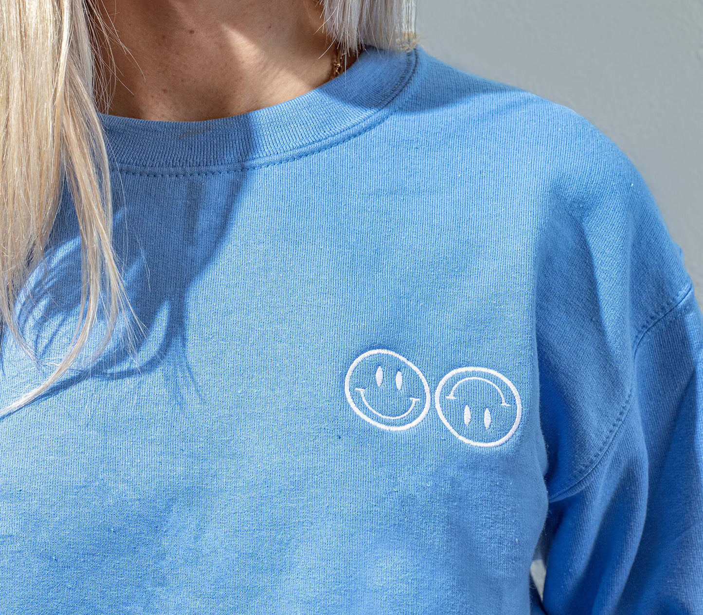 A close up of a woman wearing a blue Happy V Crewneck Sweatshirt, displaying the Happy V logo.